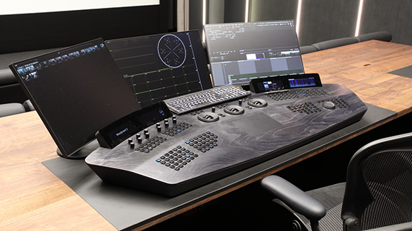 Video editing setup with three monitors and a large black wood-grain control console on a wooden desk.