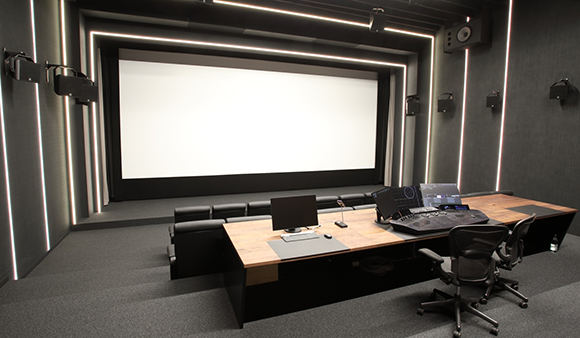 A dark screening room featuring a large blank white screen and a wooden control desk with monitors and equipment.