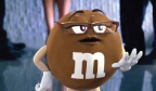 A brown M&M character with glasses and white gloves gesturing with its hands.