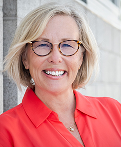 A smiling blonde woman with tortoiseshell glasses wearing a bright red collared shirt.