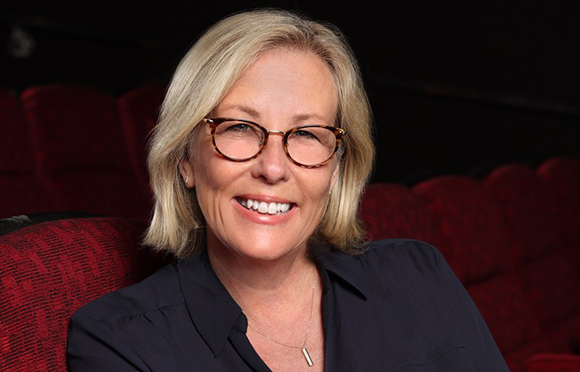 A woman with blonde hair, glasses, and a dark shirt smiles while sitting in front of red seats.