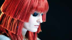 Profile of a person with vibrant red layered hair and heavy dark eyeshadow against a black background.