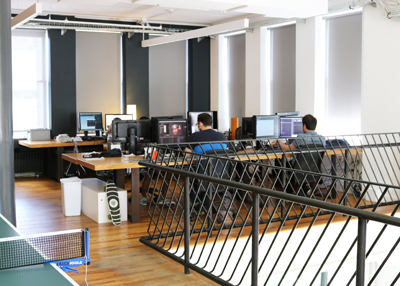 People sit at desks with computer monitors in a bright, open-plan office with large windows.