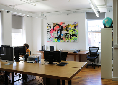 An office with wooden desks, computer monitors, and a large colorful portrait painting on a white wall.