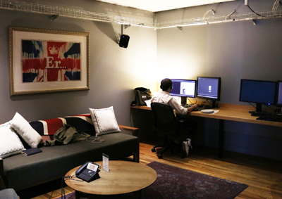 A person sits in a black office chair working on a computer at a long wooden desk in a room with a sofa.