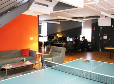 A modern office featuring a lounge with a grey sofa, a ping pong table in the foreground, and desks in the background.
