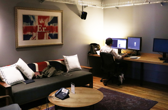 A person sits at a long wooden desk with multiple computer monitors in a room with a couch and framed art.