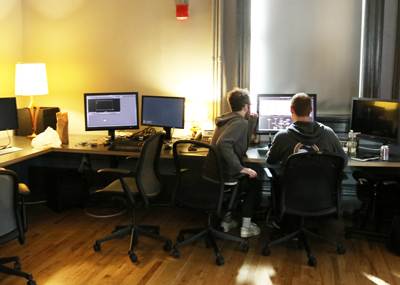 Two people sit in office chairs working at a desk with several computer monitors in a dimly lit room.