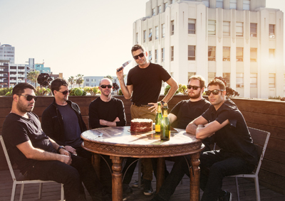 Six men in dark clothing sit around a round table on a sunny rooftop with a city building in the background.