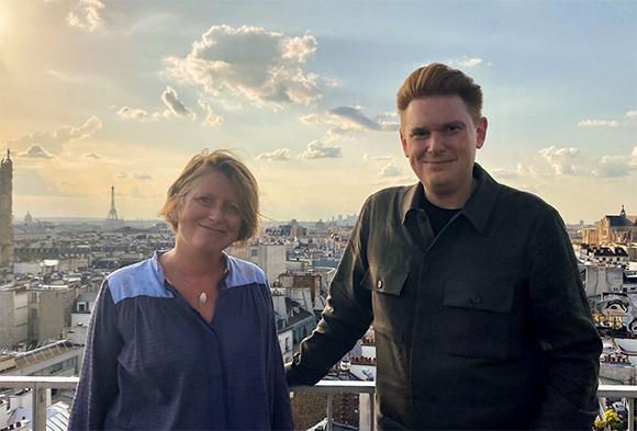 A man and a woman smile on a rooftop with the Paris skyline and Eiffel Tower in the background.