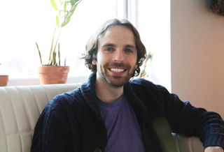 A man with medium-length brown hair and a beard smiles while sitting on a white leather sofa.