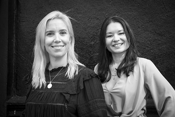 Two women smiling while standing side by side against a dark, textured wall.