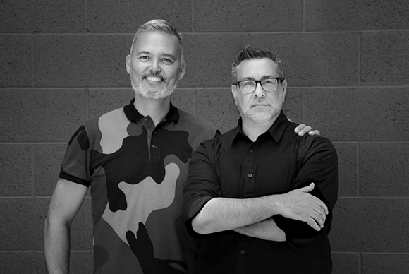 Two men standing side by side in front of a dark brick wall in a black and white portrait.