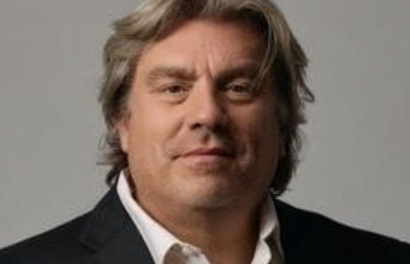 A middle-aged man with wavy brown hair wearing a black suit jacket and white shirt.
