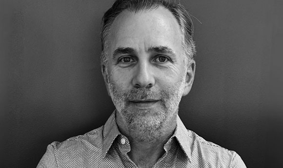 Black and white headshot of a middle-aged man with short hair and light facial stubble looking forward.