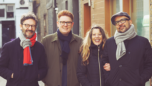 Four smiling adults in winter coats and scarves standing together on a city street.