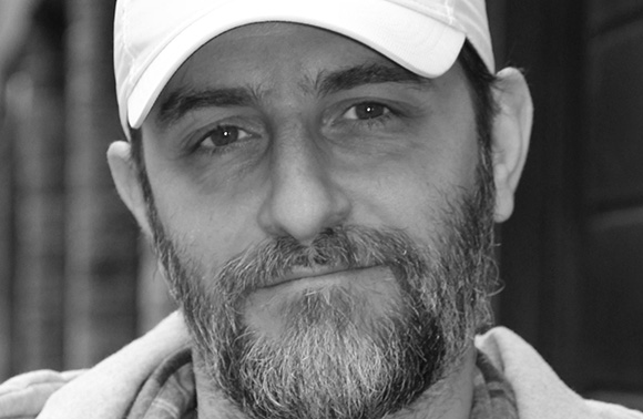 Black and white close-up of a man with a beard and a white baseball cap looking at the camera.