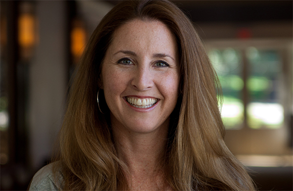 A smiling woman with long brown hair and blue eyes looking directly at the camera.
