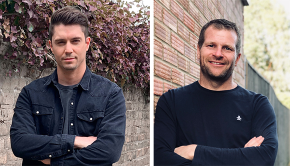 Two men with arms crossed, one in a dark denim shirt and one in a black long-sleeve shirt.
