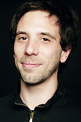 A man with dark, wavy hair and light stubble smiling against a black background.