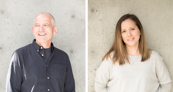 A man in a dark shirt smiling and a woman in a light sweater smiling against a concrete background.