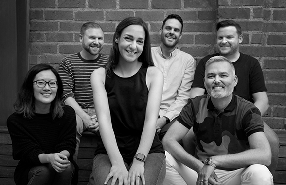 Seven diverse people smiling and posing together in a group portrait against a brick wall.
