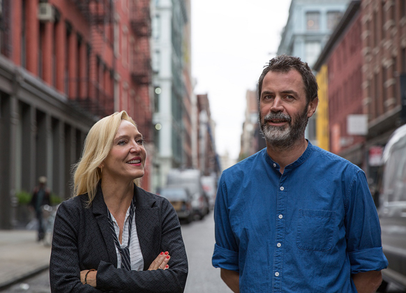 A blonde woman in a black blazer smiles at a bearded man in a blue shirt on a city street.