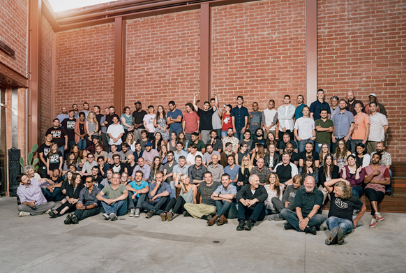 A large, diverse group of people posing together in front of a tall red brick wall outdoors.