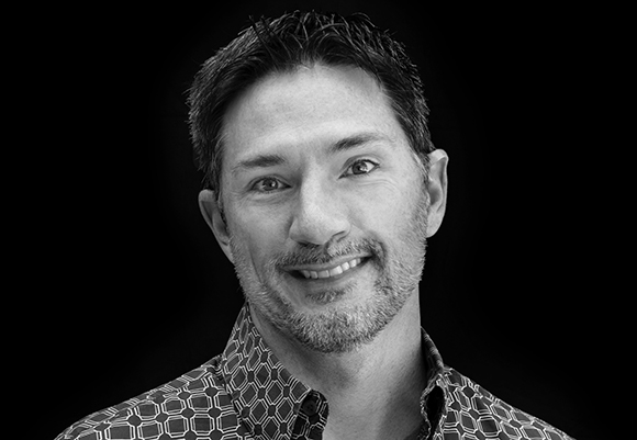 Black and white headshot of a smiling man with short dark hair and a light beard against a black background.