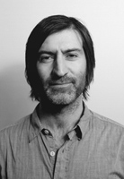 Black and white headshot of a man with medium-length dark hair and a light beard wearing a button-down shirt.