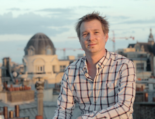 A man in a white and dark plaid shirt stands on a rooftop with a city skyline in the background.