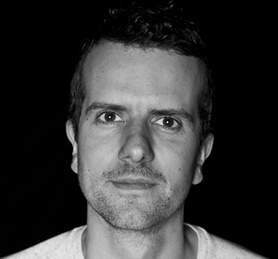 Black and white close-up portrait of a man looking directly at the camera against a dark background.