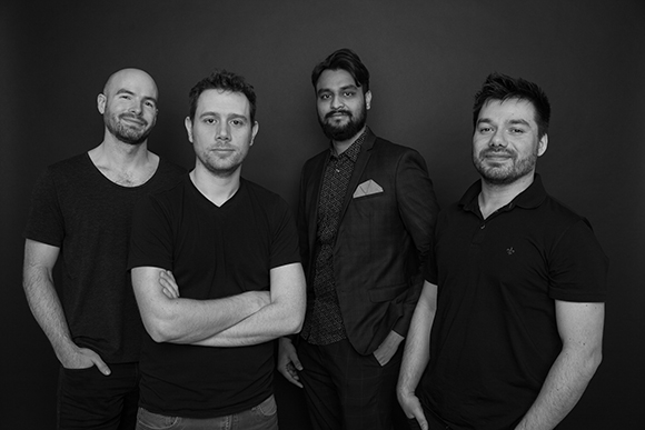 Four men standing in a row against a dark backdrop, wearing dark casual and semi-formal clothing.