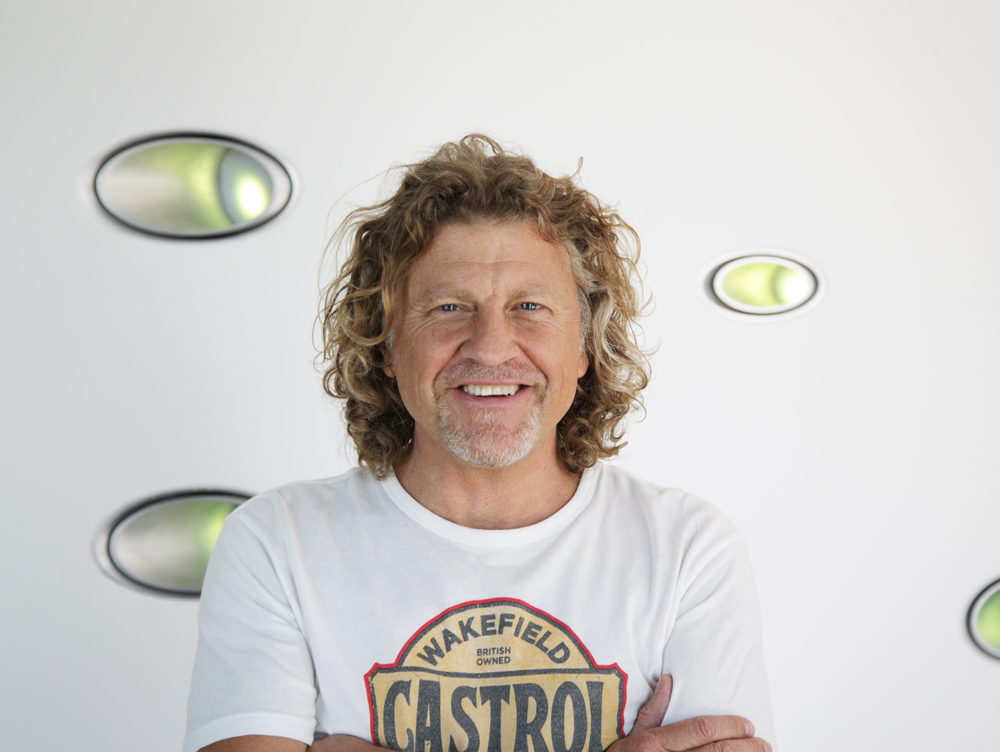 A smiling man with curly blonde hair wearing a white Castrol graphic t-shirt against a white wall.