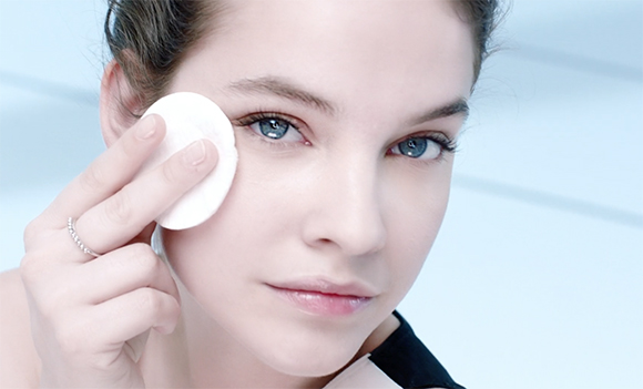 A woman uses a white cotton pad to gently wipe the skin around her eye.