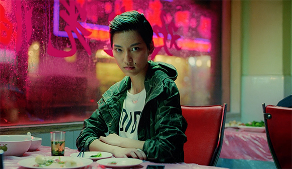 A person with short dark hair wearing a green jacket sits at a table in a restaurant with pink lighting.
