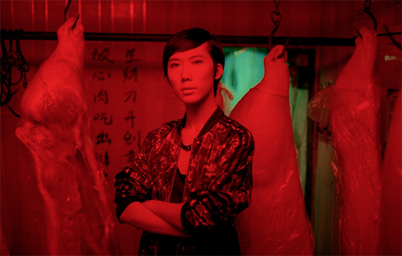 A person with short dark hair stands with arms crossed in a red-lit room with hanging meat.
