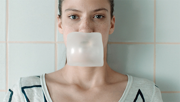 A person with freckles blowing a large, translucent white bubble of gum in front of a tiled wall.