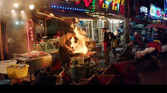 A street vendor cooks food over a large burst of flame at a busy outdoor night market stall.
