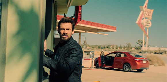 A man in a black jacket peeks from behind a teal wall at a desert gas station.