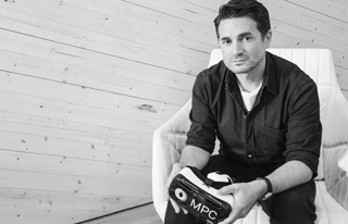 A man in a dark button-down shirt sits in a white chair holding a VR headset labeled MPC.