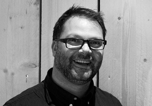 A smiling man with dark hair, a beard, and glasses poses in front of a wooden wall.