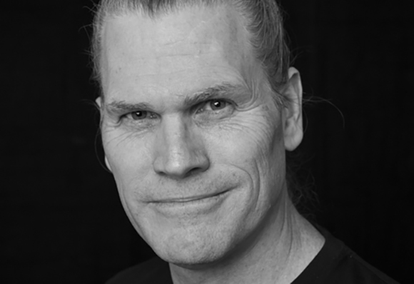 Black and white close-up portrait of a man with light hair pulled back, smiling slightly.