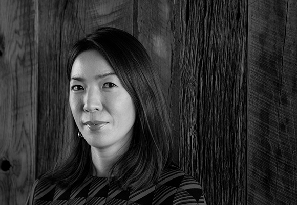 Black and white portrait of a woman with shoulder-length hair looking toward the camera against a wood background.