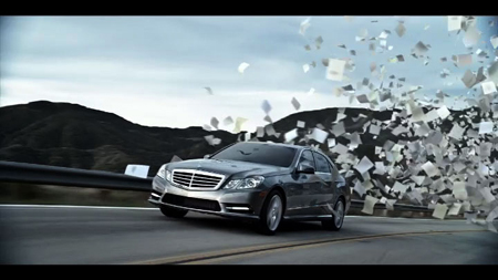 A dark luxury sedan drives along a mountain road as numerous white papers fly through the air behind it.