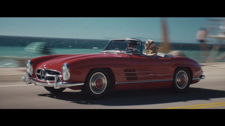 A red vintage Mercedes-Benz convertible driving on a road next to the ocean.