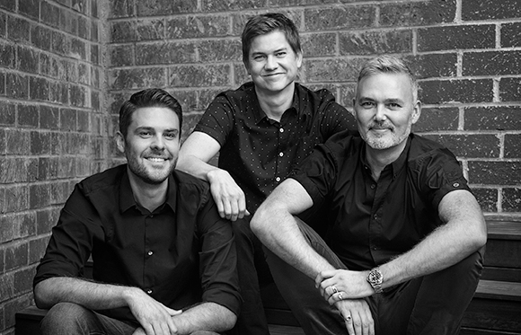 Three men in dark shirts sit together against a brick wall in a black and white portrait.