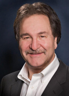 A smiling man with brown hair and a mustache wearing a dark suit and white shirt.