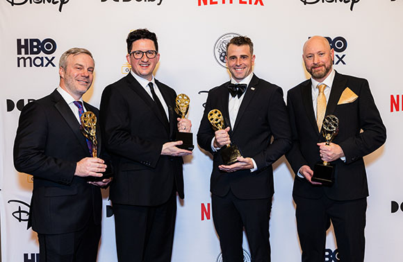 Four men in black suits and ties holding gold trophies in front of a white backdrop with logos.