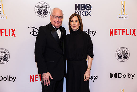 A man in a tuxedo and a woman in a black dress pose in front of a white wall with various media logos.